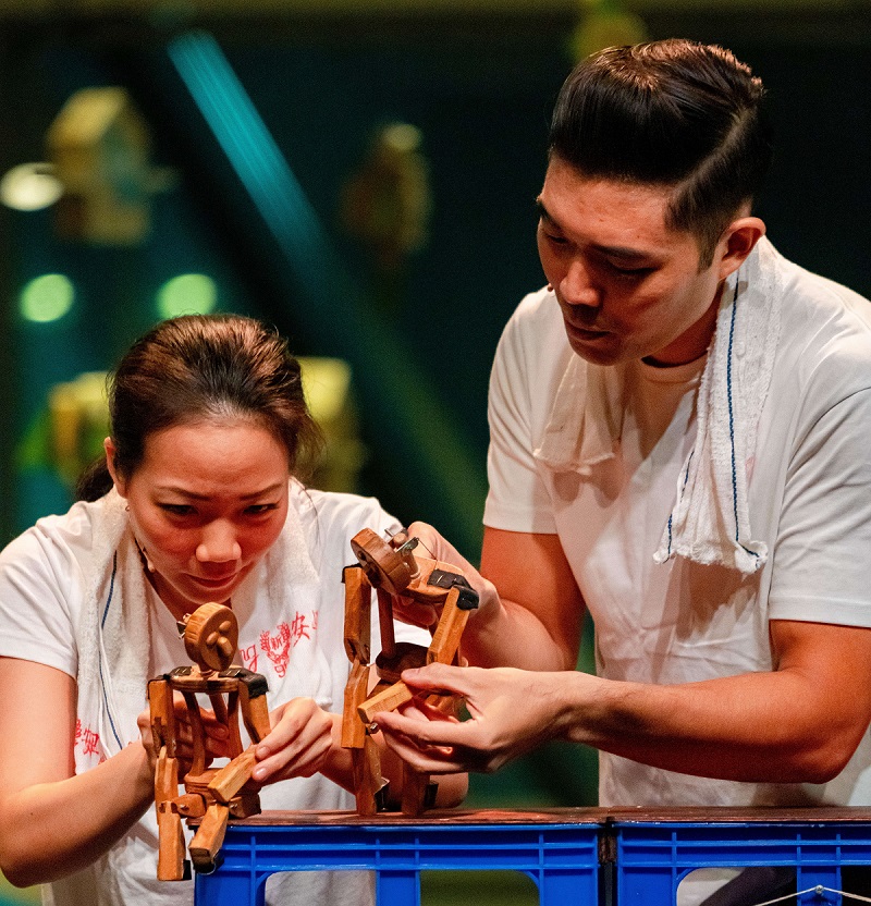 Two puppeteers in white shirts with wooden puppets on a makeshift stage made of blue crates.